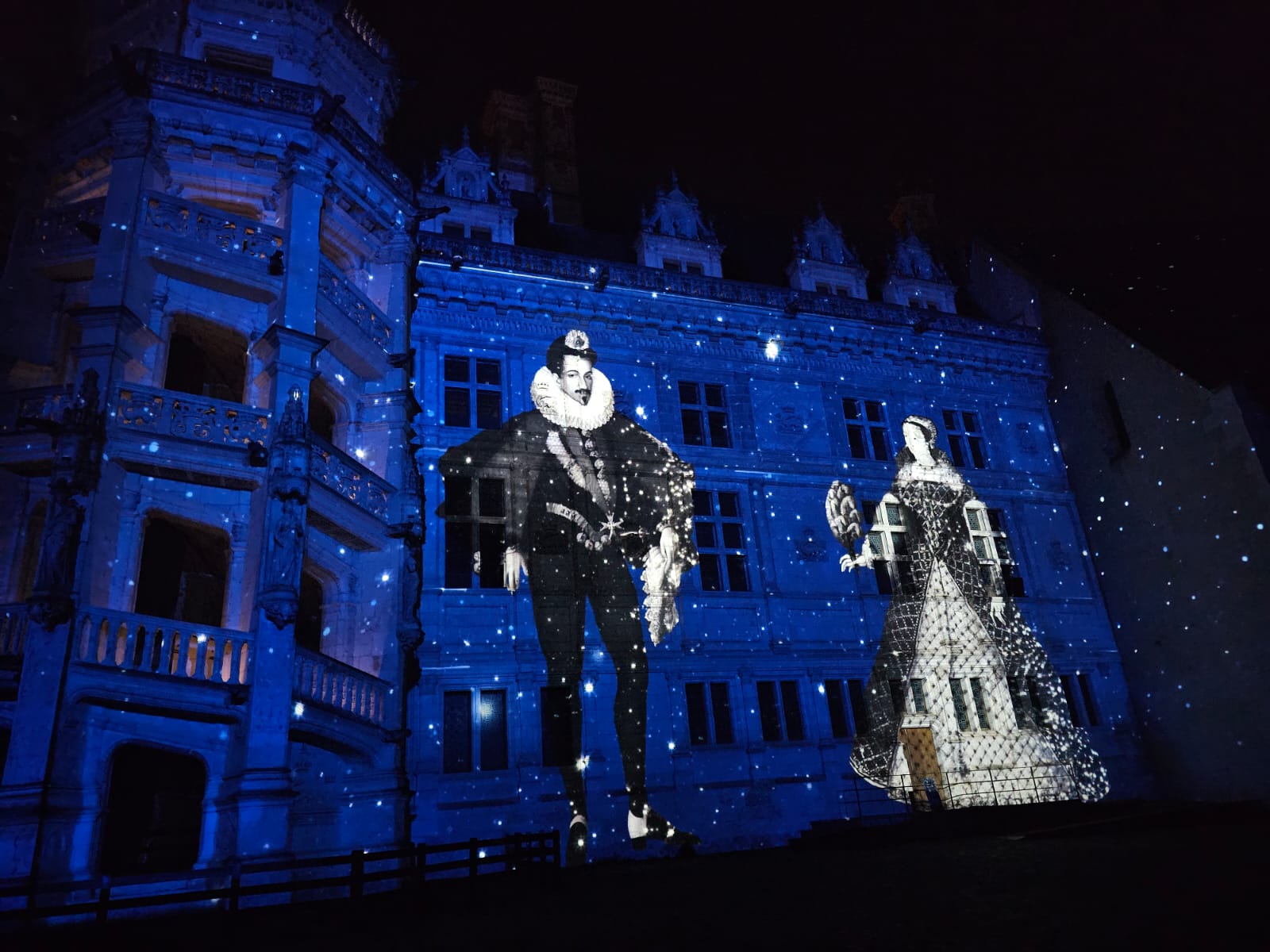 Projection nocturne sur la façade du château de Blois : deux personnages historiques en costume Renaissance apparaissent en lumière sur les murs bleus du monument.